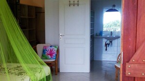 Una habitación con una cama cubierta por una tela mosquitera verde, una puerta blanca, una silla con un cojín de colores y vistas a un baño con un lavabo y un espejo.