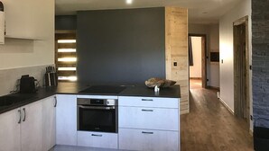 A modern kitchen with a built-in oven, a countertop with a stove, and a wooden accent wall. - Chalet Le Lynx (Ferdrupt)