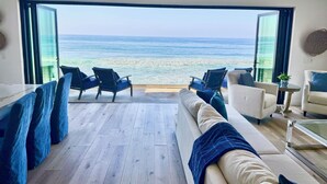 Un moderno salón con un amplio ventanal que da a una vista de la playa. Hay dos sillones azules y un sofá blanco con una manta azul.