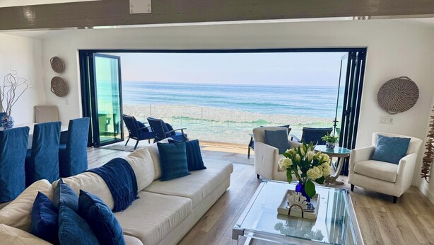 Una sala de estar moderna con un sofá grande, sillones y una mesa de centro de vidrio. Un ventanal ofrece vistas a la playa.