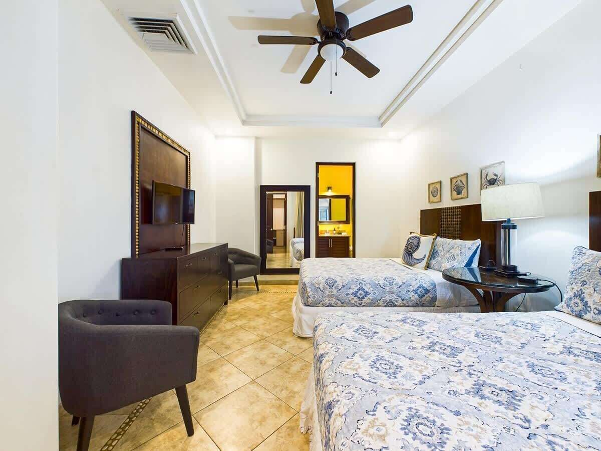A hotel room with two beds, a desk, a chair, a ceiling fan, and a TV.
