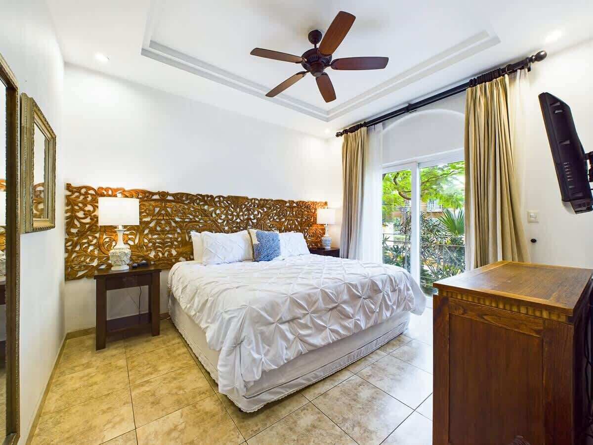 A bedroom with a bed, a nightstand, a ceiling fan, a wooden headboard, and a view of greenery outside.
