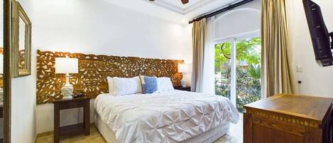 A bedroom with a bed, a nightstand, a ceiling fan, a wooden headboard, and a view of greenery outside.