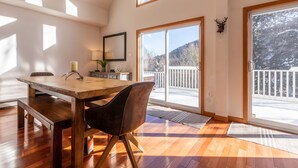 A dining area with a wooden table and chairs, a lamp, and a view of a snowy landscape through large windows. - Quiet Vermont Winter Getaway (Bridgewater)