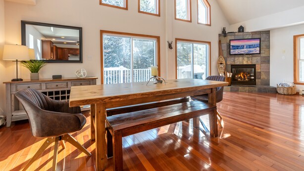 A spacious living room with a large wooden dining table, comfortable seating, a fireplace, and large windows offering a view of the outdoors. - Quiet Vermont Winter Getaway (Bridgewater)