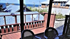 Una terraza de madera con dos sillas, vista al mar y un edificio con techo inclinado al fondo.