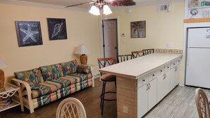 Uma cozinha compacta com uma ilha central, uma sala de jantar com sofá e um ventilador de teto.