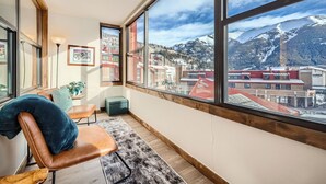 A room with large windows offering a view of a mountain and buildings. There is a wooden bench, a small table with a plant, and a rug on the floor. - Remodeled unit & mountain view in the ♥ of Central Village, walk to the lifts!  (Frisco)