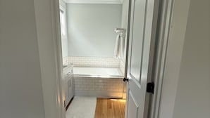 Un couloir étroit mène à une salle de bain équipée d’une baignoire, d’un mur recouvert de carreaux blancs et d’un plancher en bois.