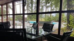 Una taula amb superfície de vidre i cadires en una habitació amb finestrals grans que donen a una piscina i vegetació.