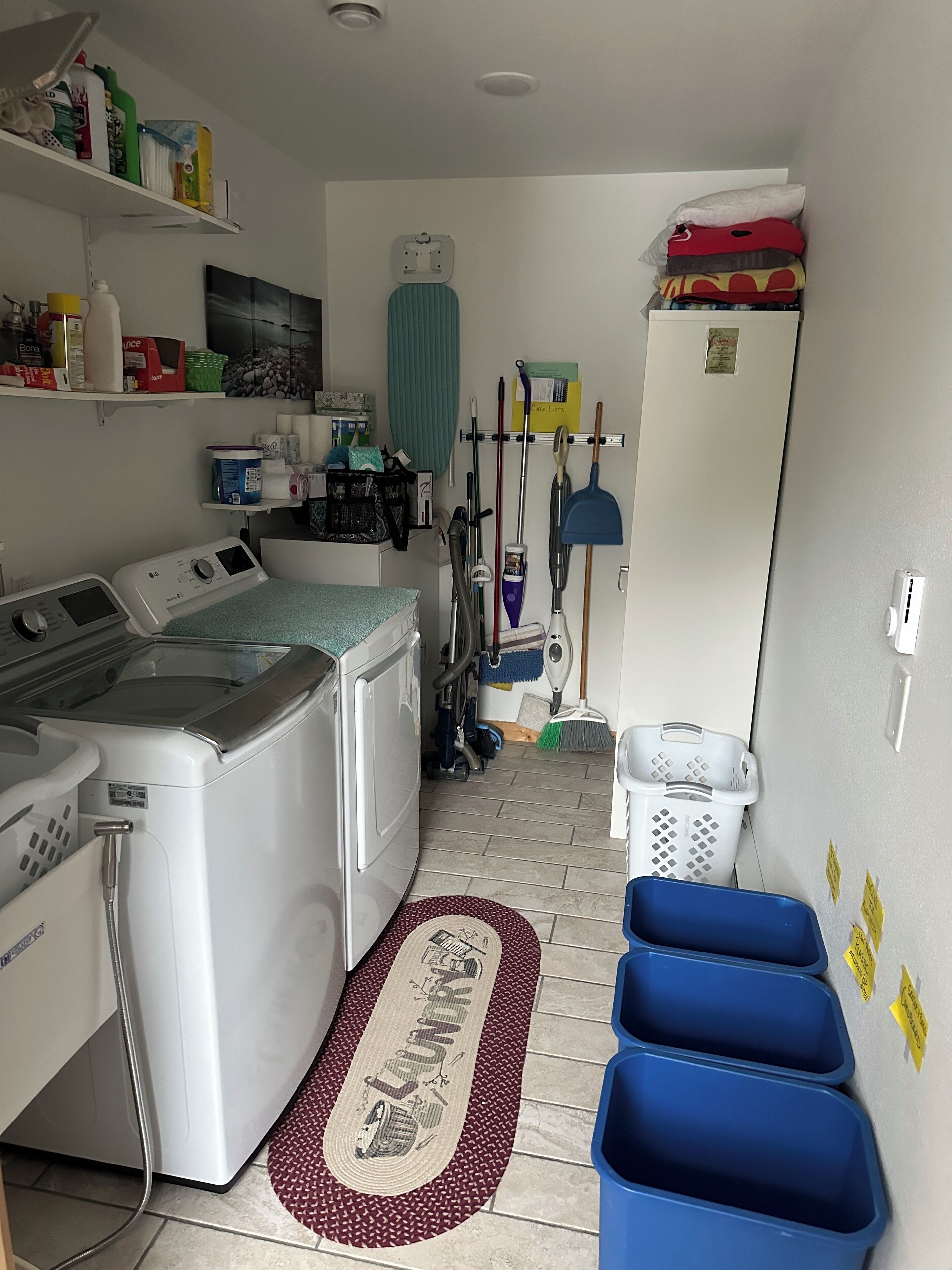 A laundry room with a washing machine, dryer, and various cleaning supplies on shelves. There are blue storage bins and a mop on the floor.