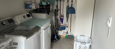 A laundry room with a washing machine, dryer, and various cleaning supplies on shelves. There are blue storage bins and a mop on the floor.
