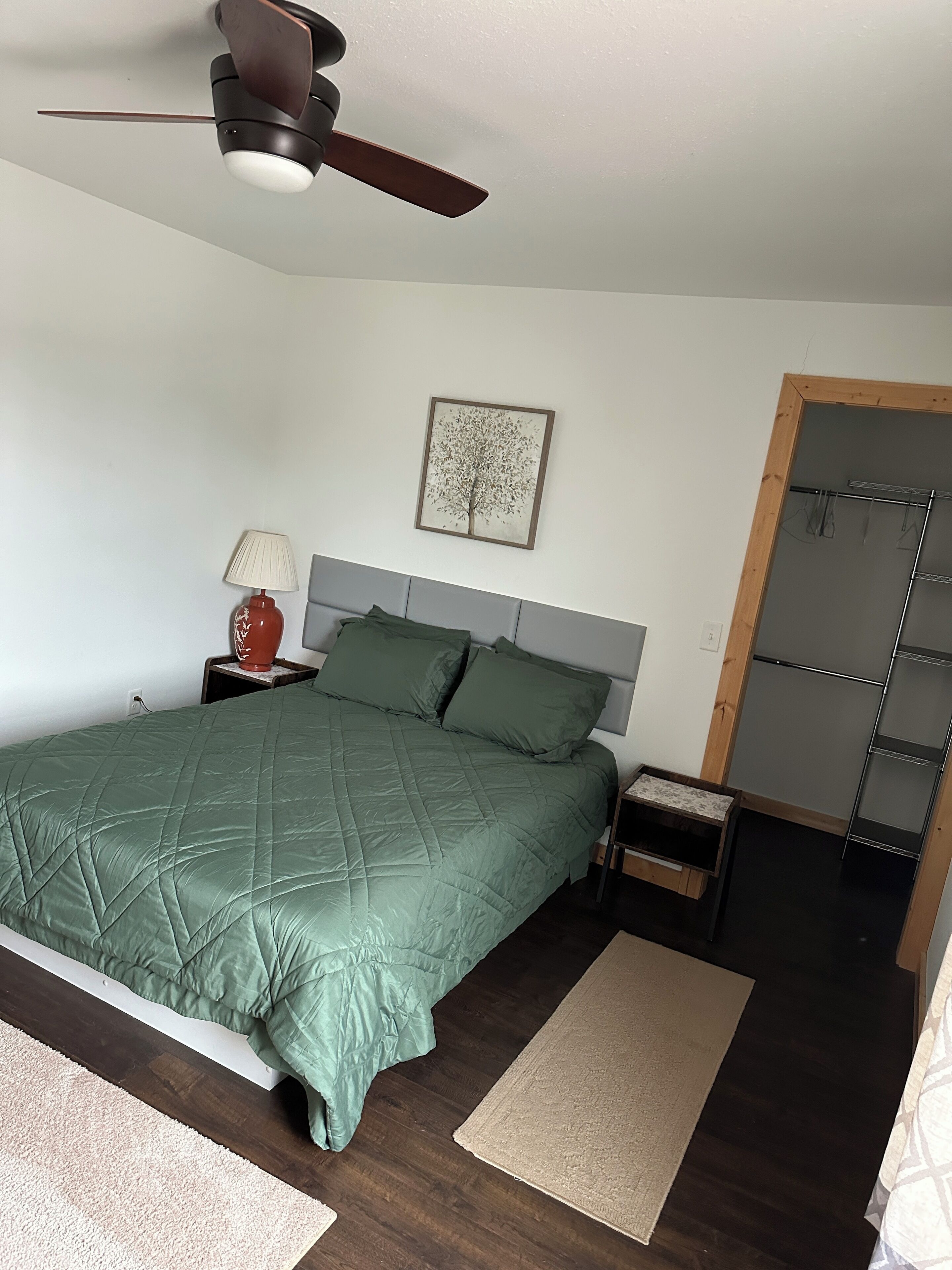 Une chambre à coucher comprenant un lit, une table de chevet, une lampe, un ventilateur de plafond, un tableau encadré, une armoire et une porte.