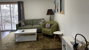 Una sala de estar con un sofá verde, una mesa de centro blanca, un tapete estampado y una mesita auxiliar blanca.