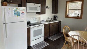 Cocina compacta con armarios de madera oscura, un refrigerador blanco, un microondas, una estufa y un lavabo. Hay una ventana con vista a árboles y un ventanal con marco de madera sobre el lavabo.