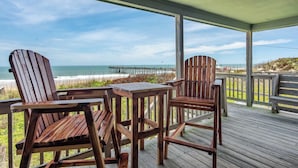 Terraza de madera con sillas Adirondack y una mesa, con vista a una playa y un muelle.