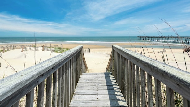 Una pasarela de madera que conduce a una playa arenosa con un muelle a lo lejos.