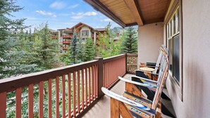 A balcony with a wooden railing, a table with magazines, and a view of a mountain resort with multiple buildings and trees. - Ski-In/Ski-Out Corner Unit 2BR/2BA Mountain Views & In-Unit Laundry (Salt Lake City)