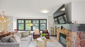 A modern living room with a stone fireplace, a flat-screen TV, and a dining area with wooden furniture. - Ski-In/Ski-Out Corner Unit 2BR/2BA Mountain Views & In-Unit Laundry (Salt Lake City)