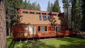 En tvåvånings timmerstuga med en veranda, omgiven av höga träd.