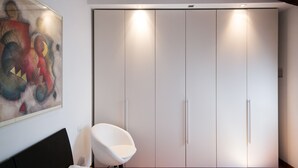 A bedroom with a bed, a white chair, a wardrobe, and a painting on the wall.