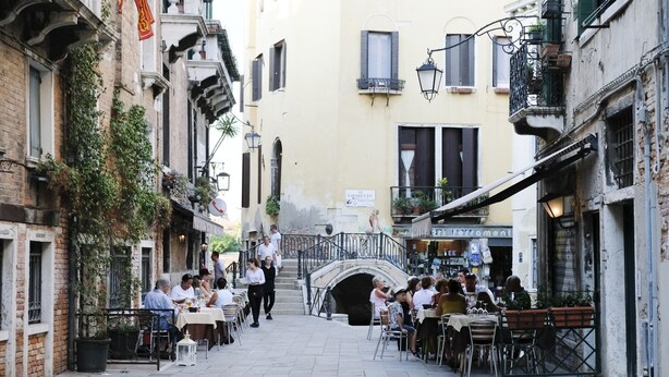 A narrow street with outdoor seating, a bridge, and buildings on either side.