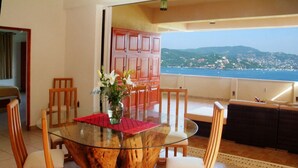 A dining area with a glass table surrounded by wooden chairs, a vase with flowers, and a view of the sea and hills in the background.