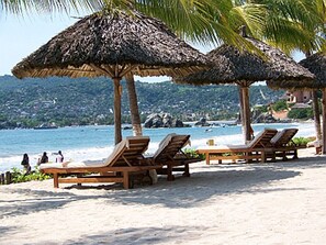 A beach with wooden lounge chairs and thatched umbrellas, overlooking a coastal area with buildings in the distance.