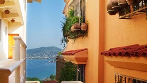 A narrow outdoor corridor with a view of the sea, a balcony with potted plants, and a staircase leading to a higher level.