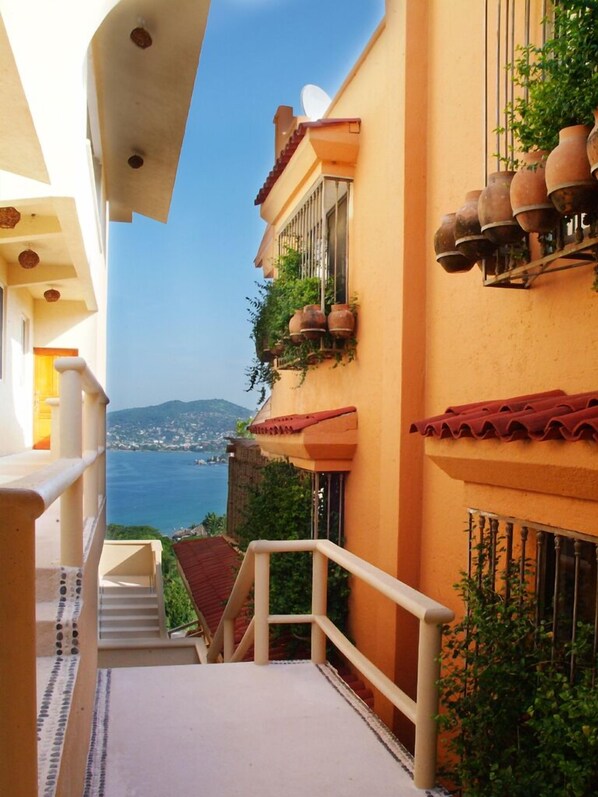 A narrow outdoor corridor with a view of the sea, a balcony with potted plants, and a staircase leading to a higher level.