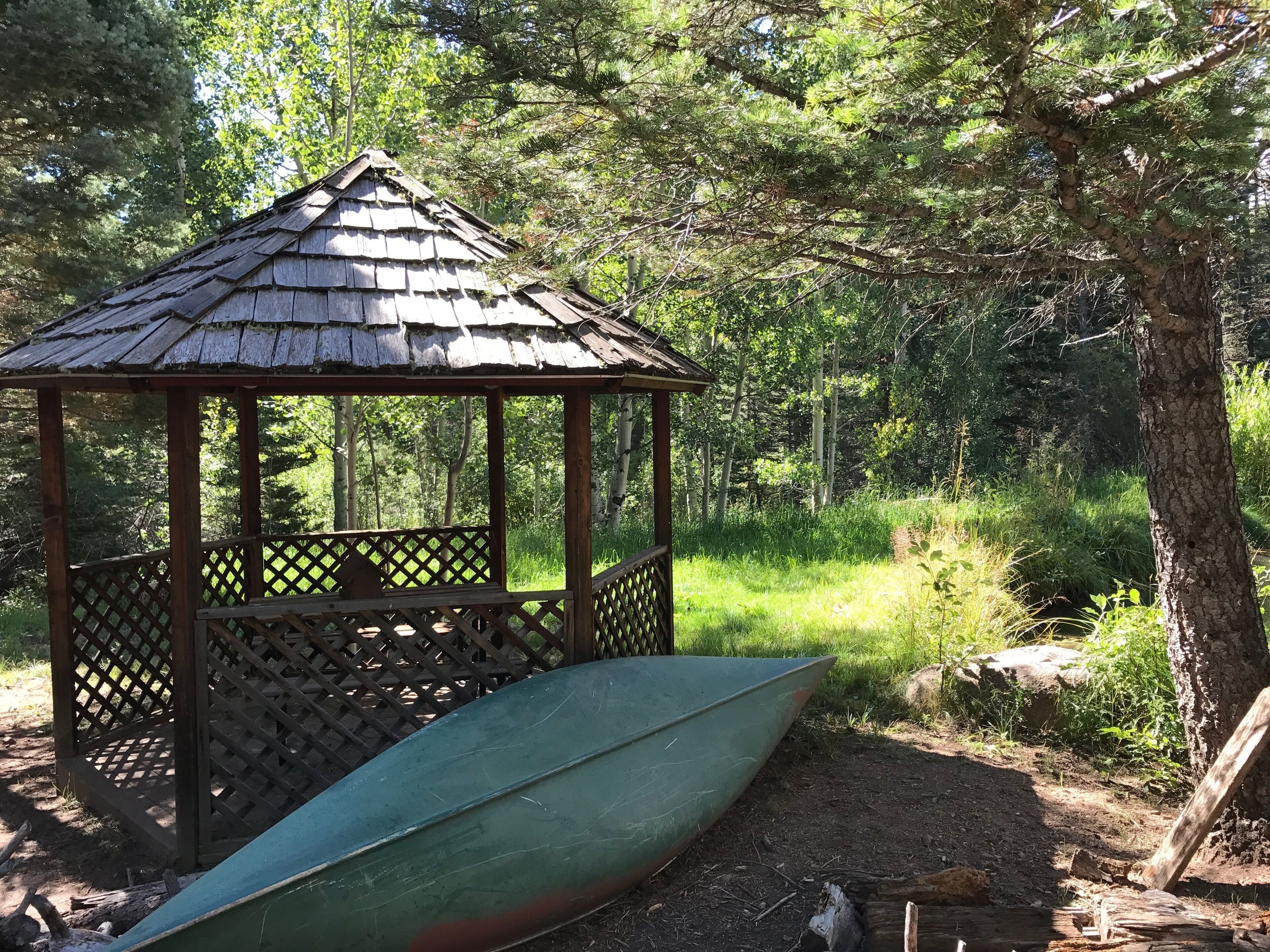 Un belvédère en bois à motifs de treillis, entouré d’une végétation luxuriante et d’arbres.