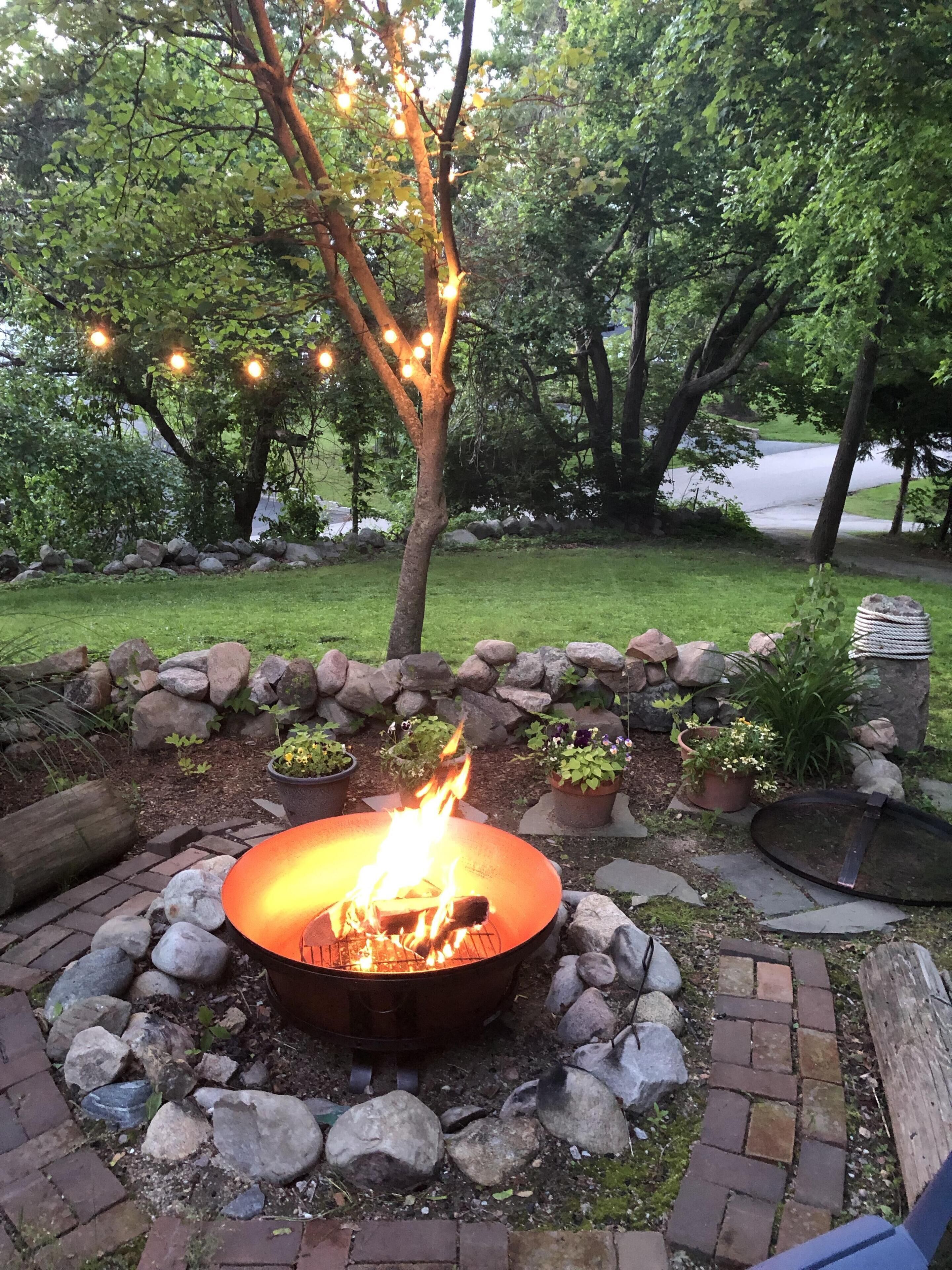 A fire pit surrounded by rocks and potted plants, with a tree overhead and string lights.