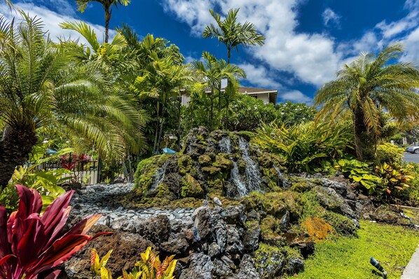 A tropical garden with a waterfall, lush greenery, and a building in the background.