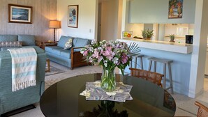 A living room with a sofa, a coffee table with a vase of flowers, a ceiling fan, and a kitchen area with a counter and stools. - Wailea Ekahi #45-E Ocean View Beachfront Resort Great Rates, Pool, Near Beach (Wailea)