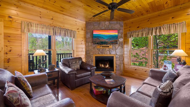 A cozy cabin interior with wooden walls, a stone fireplace, leather sofas, and a ceiling fan. - Gatlinburg, Hot Tub, Huge Arcade, Dual Racing, Theater, Walk-2-Pool, Free Tix (Gatlinburg)