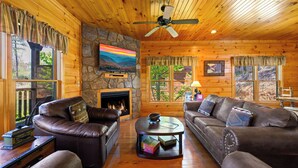 A cozy log cabin interior with leather sofas, a stone fireplace, a television, and a stone wall. - Gatlinburg, Hot Tub, Huge Arcade, Dual Racing, Theater, Walk-2-Pool, Free Tix (Gatlinburg)