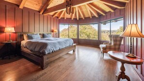 Een slaapkamer met een houten plafond, een groot bed, een stoel, een tafel en uitzicht op het groen.