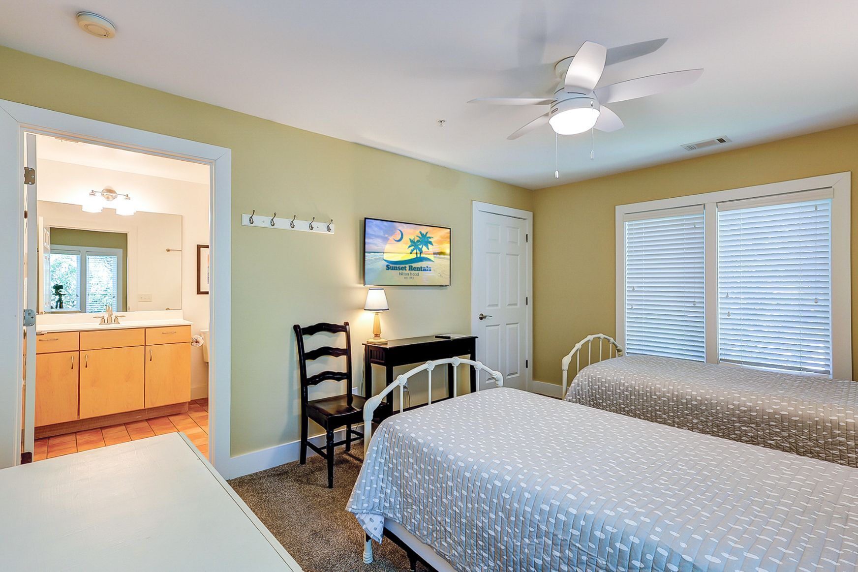 A hotel room with two beds, a desk, a chair, a small table, a ceiling fan, and a bathroom visible through an open door.