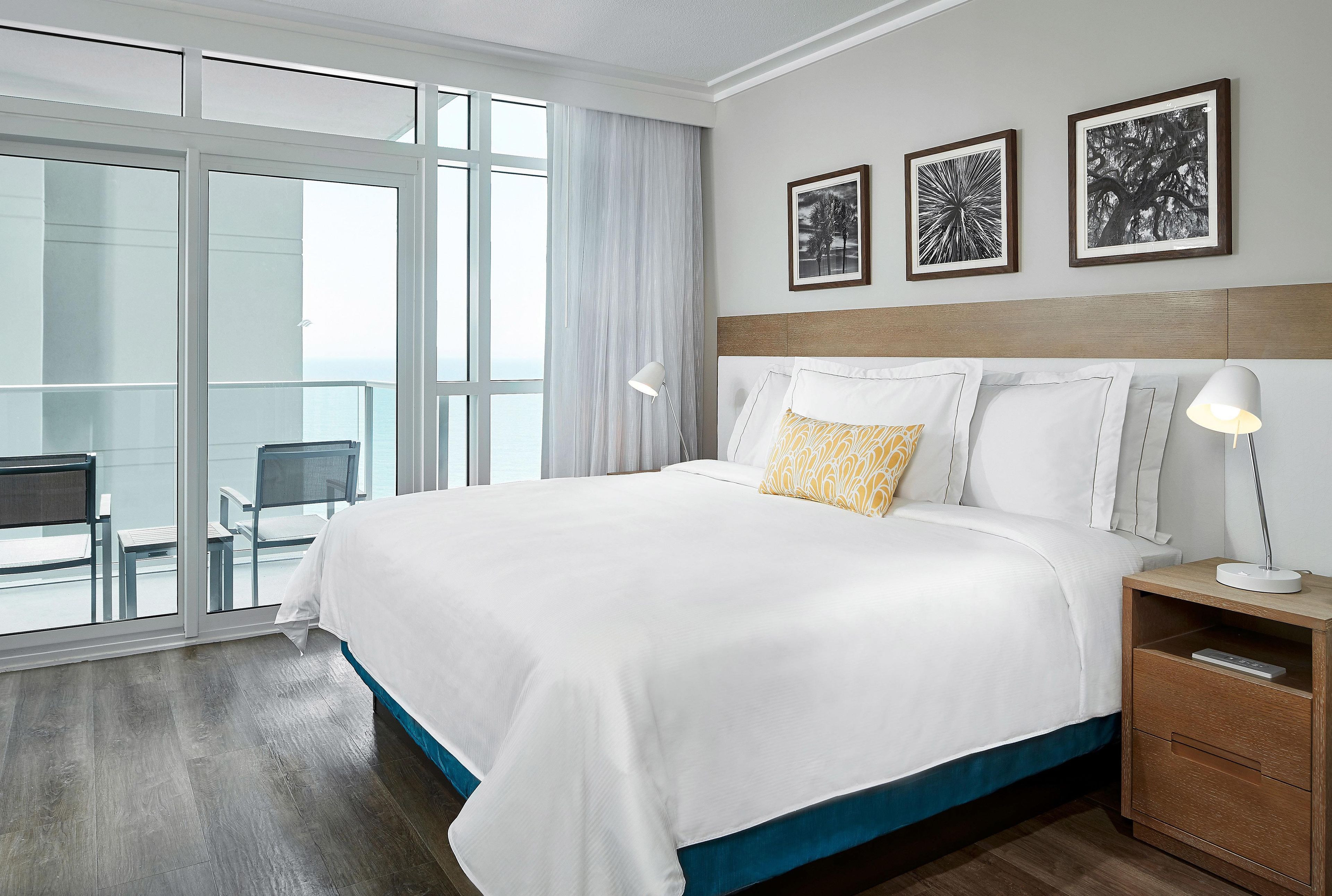 A hotel room with a large bed, a nightstand, a lamp, and a view of the outdoors.