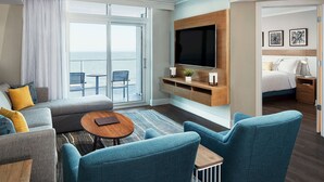 A modern hotel room with a large window, a sofa, two armchairs, a small table, a TV mounted on the wall, and a bed with a bedside table.