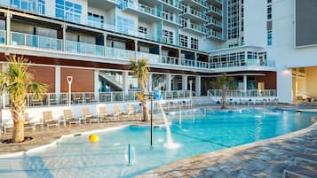 A swimming pool with lounge chairs and palm trees, surrounded by a multi-story building with balconies.