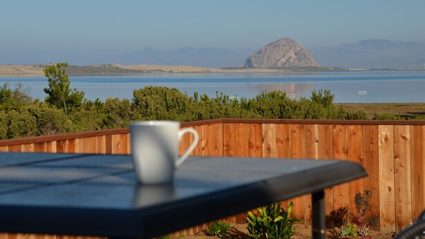 Een houten terras met een tafel en een witte mok, met uitzicht op een meer en verre bergen, en een houten omheining.