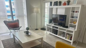 A living room with a white coffee table, yellow sofa, and a TV on a white stand. - Sunrise+Sunset on the Beach  (Hollywood)