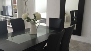 A modern dining area with a glass-top table, black chairs, a white vase with flowers, and a large mirror on the wall. - Sunrise+Sunset on the Beach  (Hollywood)