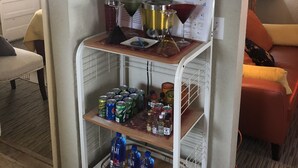 Ett kompakt bostadsutrymme med ett litet köksutrymme som inkluderar ett vinställ, en hylla med olika flaskor, en bardisk med glas och en nedre hylla med burkdrycker och flaskvatten.