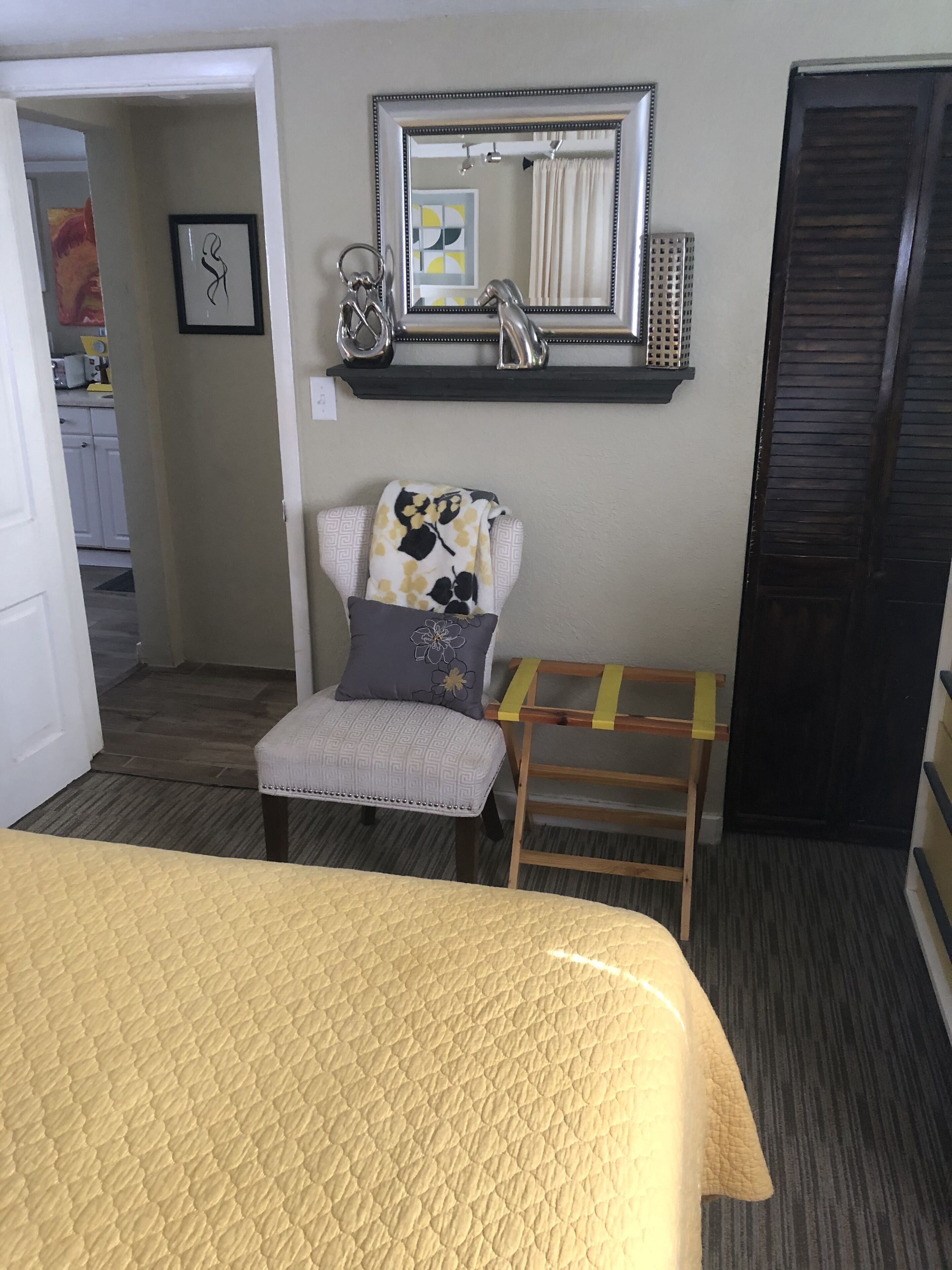 A hotel room with a bed, a chair with a cushion, a small table, a mirror, and a door.