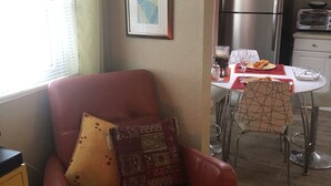 Ett vardagsrum med en röd läderfåtölj, ett matbord med stolar, en köksdel med vitvaror och en inramad tavla på väggen.