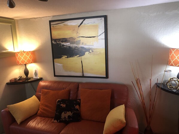 A living room with a brown leather sofa, two lamps with patterned lampshades, a framed abstract painting, and a vase with reed sticks.