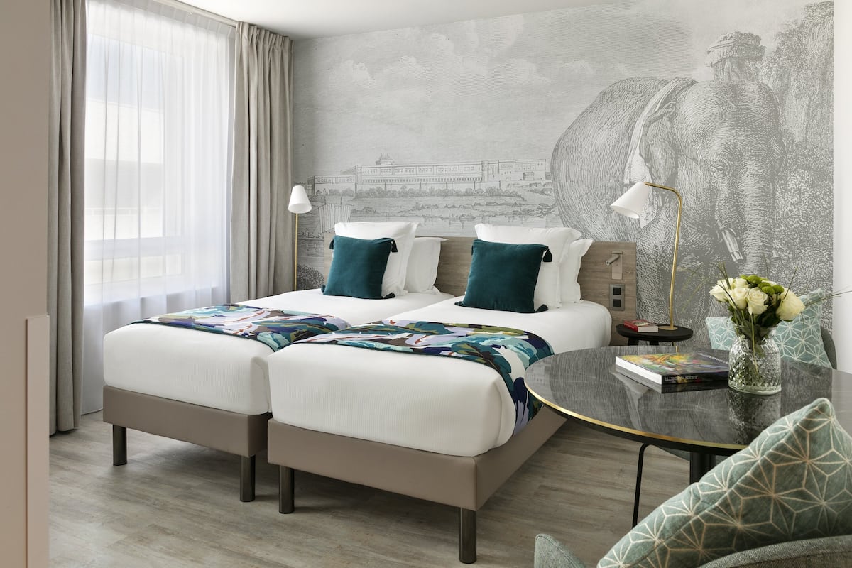 A hotel room with a bed, two bedside tables, a lamp, a vase with flowers, and a mural on the wall.
