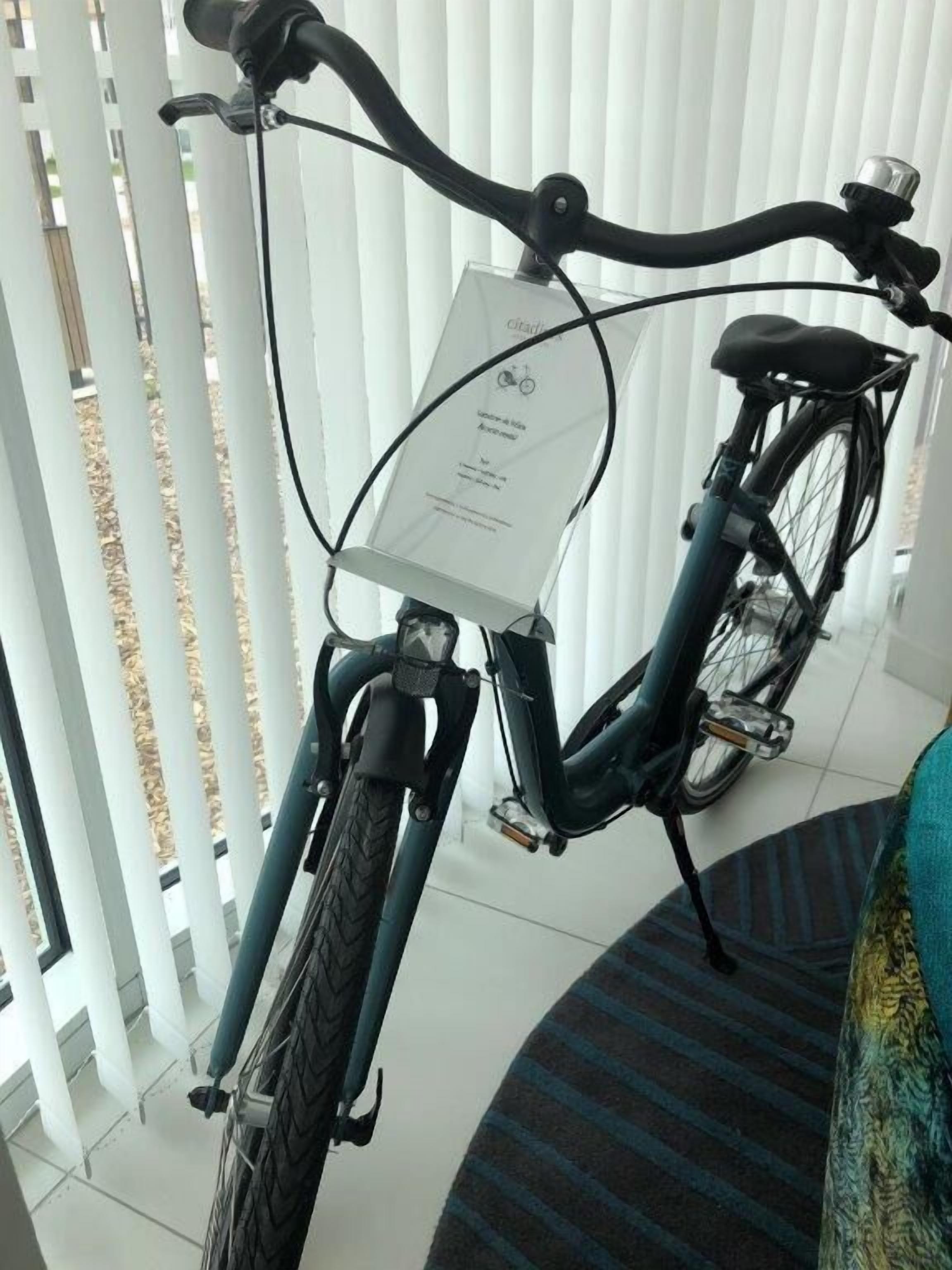 Una bicicleta con una etiqueta blanca pegada al frente, estacionada adentro cerca de un pilar blanco y un tapete estampado.